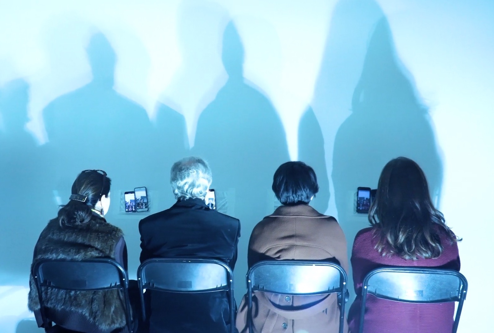 Four audience members stay seated facing the wall looking at their image on their phones mounted on the wall in a clear sleeve. The lighting produces four shadows along the wall.